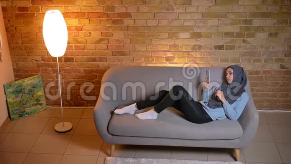 在舒适的家中年轻迷人的穆斯林女性在沙发上放松手机短信的特写镜头视频的预览图