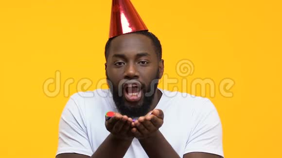 有趣的非洲裔美国男性戴着派对帽把纸屑吹进镜头生日视频的预览图