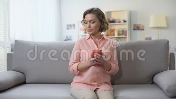 悲伤的年轻女性拿着一杯茶坐在沙发上看着相机怀念视频的预览图