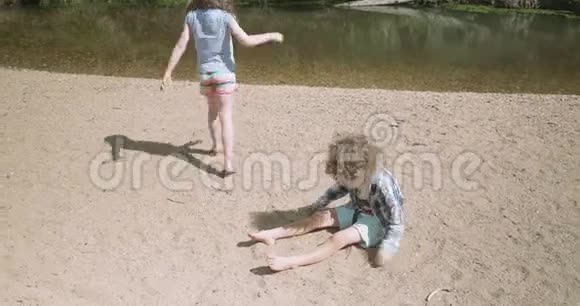 小男孩坐在河滩上用沙子盖住自己小女孩走过视频的预览图