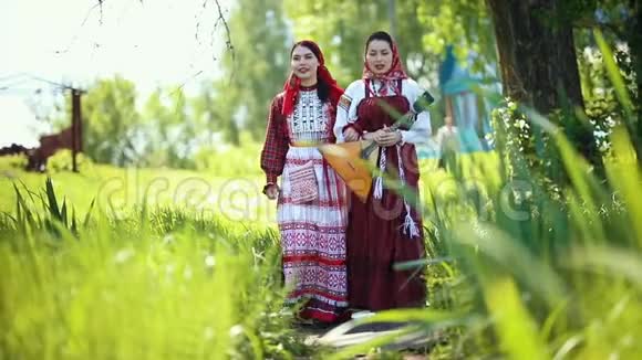 两个穿着俄罗斯传统服装的年轻女子走在田野上唱歌其中一个拿着巴拉莱卡视频的预览图
