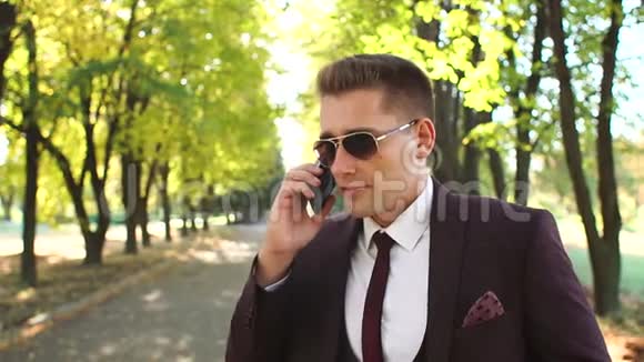 戴着太阳镜的商人在秋天公园散步时打电话视频的预览图