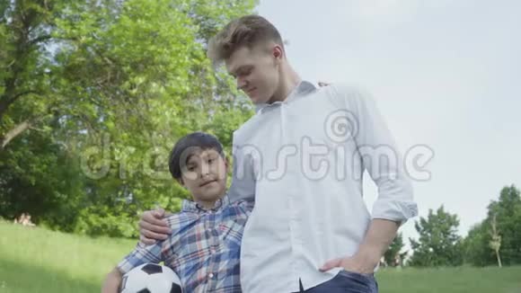 可爱的孩子拿着足球拥抱爸爸在公园里看镜头家庭休闲户外活动那个男孩视频的预览图