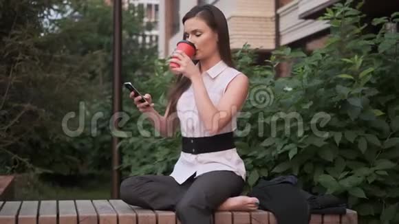 赤脚坐在公园长凳上休息的女商人年轻女子喝咖啡用手机聊天视频的预览图