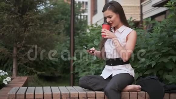 一位商务女性坐在公园长椅上休息的肖像女孩喝咖啡用手机视频的预览图