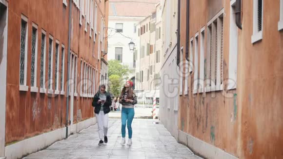 两个穿暖和衣服的年轻女旅行者走在狭窄的街道上交谈视频的预览图