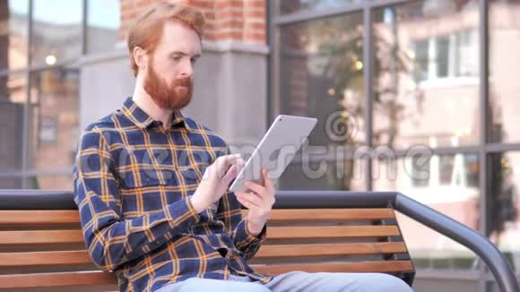 红胡子青年坐在长凳上用平板电脑视频的预览图