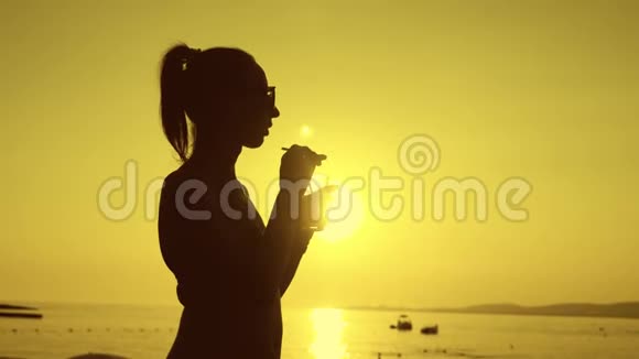 剪影年轻女子在日落时在海滩上喝鸡尾酒视频的预览图