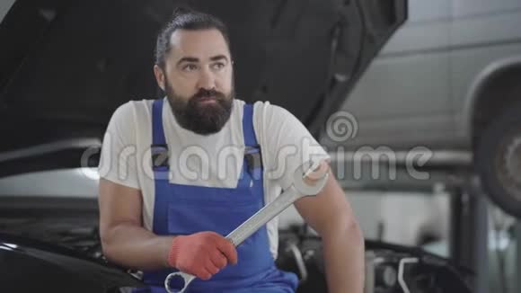 一张英俊的留着胡子的机械师的肖像扳手坐在汽车修理店里一辆破车的引擎盖上视频的预览图