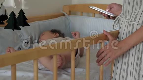 孩子在婴儿床里玩手机妈妈站在床边用电话视频的预览图