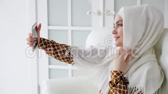 年轻的穆斯林妇女戴着头巾坐在沙发上的智能手机上自拍视频的预览图