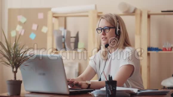 一个年轻女孩微笑着在耳机上交谈并解释了坐在办公桌前的重要工作部件视频的预览图