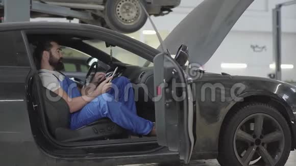 成功的机械师坐在车里在他的手机上验证信息穿制服的胡子男视频的预览图