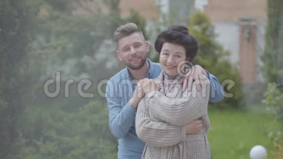 幸福成熟的女人站在花园里的大房子前面成年的孙子抱着她把手放在她身上视频的预览图
