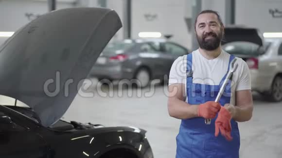 成年机械师站在汽车附近手里拿着一把巨大的扳手穿制服的胡子男视频的预览图