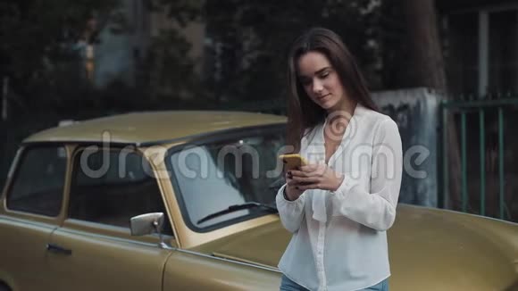 年轻迷人的布鲁内特白种人女孩站在老时尚的金色汽车边使用她的智能手机和视频的预览图