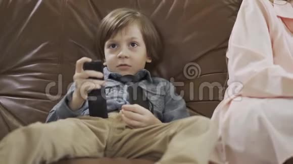 肖像小可爱男孩坐在皮革沙发上双腿分开用遥控器在电视上更换频道可爱的孩子视频的预览图
