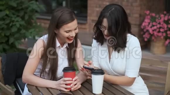 年轻女子在电话里给她的朋友看了些东西两个女商人坐在一家夏日咖啡馆的桌子旁视频的预览图