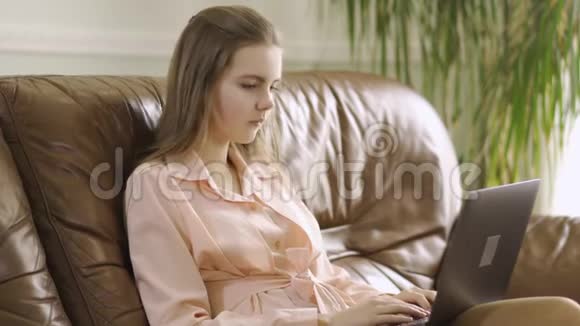 肖像可爱集中的年轻女孩坐在皮革沙发上打字在笔记本电脑键盘上现代的休闲视频的预览图