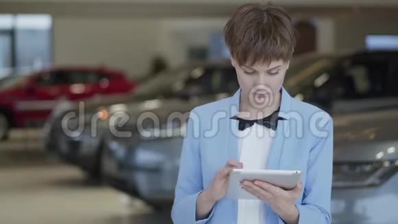 在车展上自信的漂亮女人穿着正式服装在模糊的汽车前站着她的平板电脑夫人视频的预览图