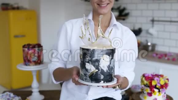 穿着白色衬衫的女性糖果师在厨房里呈现现代装饰的黑白蛋糕黄色冰箱和视频的预览图