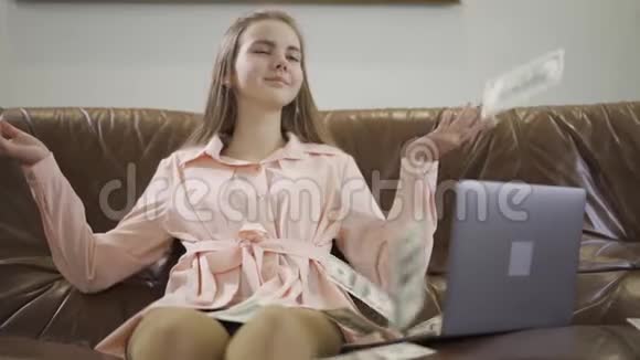 漂亮的少女坐在笔记本电脑前的皮革沙发上美元落在女孩身上孩子微笑着视频的预览图
