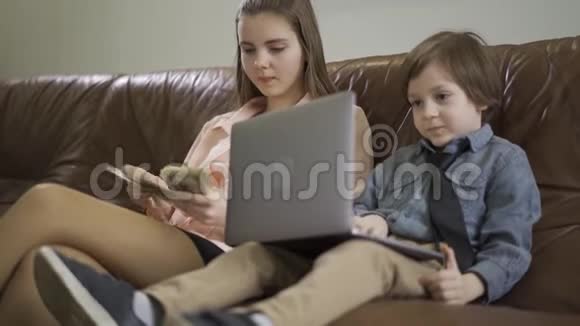 肖像姐姐和弟弟坐在皮革沙发上那个拿着笔记本电脑的男孩那个正在数数的女孩视频的预览图