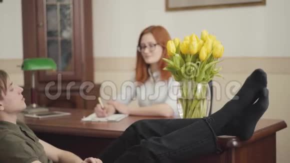 年轻人坐在一位心理学家的办公室里脚踩在桌子上告诉她他的问题女性视频的预览图