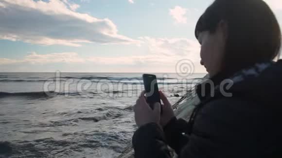 年轻女孩站在海边看着智能手机视频的预览图