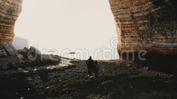在史诗般巨大的岩石拱门诺曼底下幸福的游客男女沿着令人惊叹的卵石海滩漫步的空中后景视频的预览图