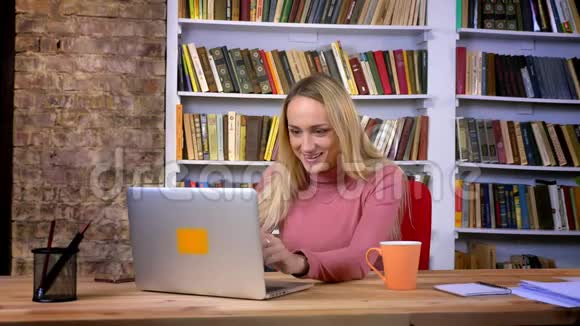 集中的高加索女孩的肖像穿着令人满意的工作与笔记本电脑在书架背景视频的预览图