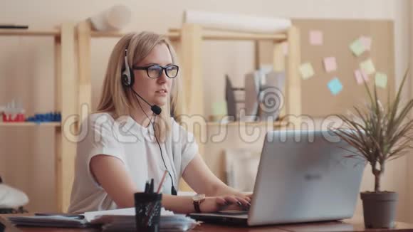 戴着眼镜和白衬衫的女孩白种人在办公室工作同时在耳机和耳机上交谈视频的预览图
