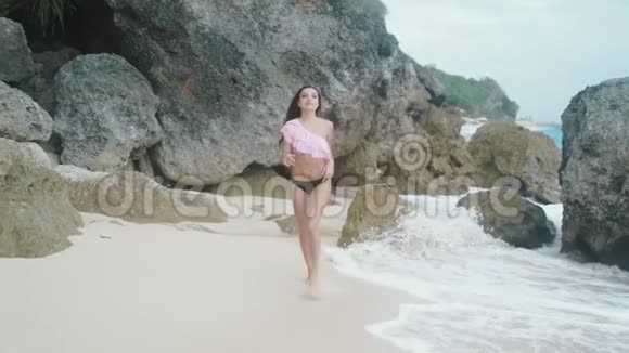 慢动作穿着泳装的女孩带着白沙和巨浪美丽的岩石和石头沿着海滩奔跑视频的预览图