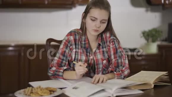 漂亮的十几岁女孩坐在家里的桌子上做作业女学生把课文从书上改写成视频的预览图