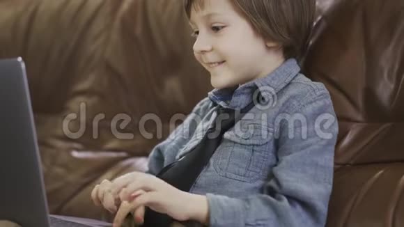 肖像可爱的小男孩穿着牛仔裤夹克坐在皮革沙发上玩着笔记本电脑开心地笑着这就是视频的预览图