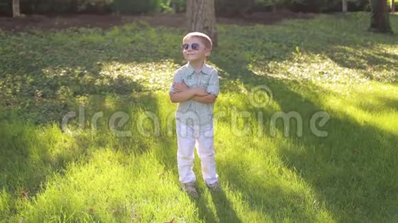 穿着白色裤子衬衫和太阳镜的可爱小男孩站在绿草上视频的预览图