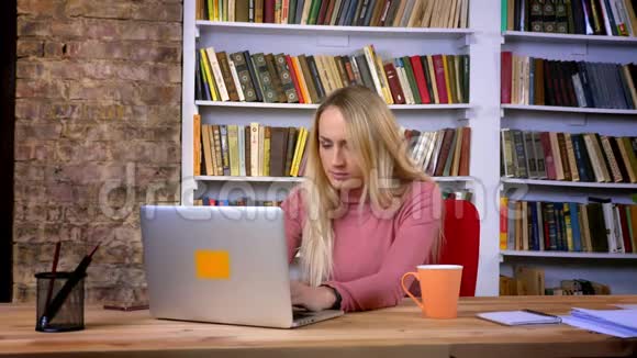 无聊的白种人女孩的肖像穿着笔记本电脑工作在书架背景上打哈欠视频的预览图