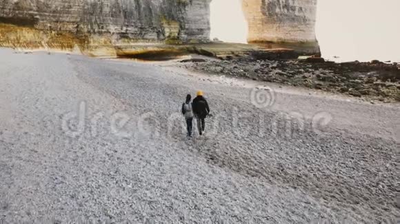 无人机镜头跟随幸福浪漫的旅游夫妇沿着令人惊叹的卵石海滩漫步到诺曼底的史诗般的白色岩石视频的预览图