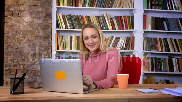 白种人年轻女孩的肖像穿着刺坐在笔记本电脑前害羞地摆姿势在书架上微笑视频的预览图