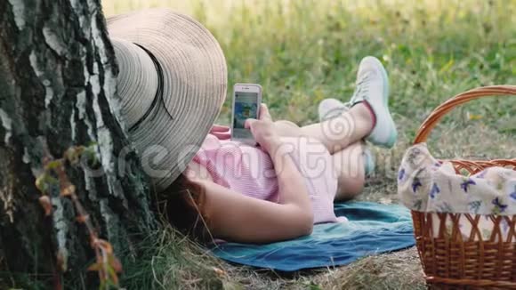野餐时间快乐的小女孩戴着圆帽子躺在树边用着手机女子网上冲浪视频的预览图