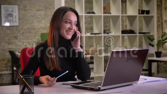 年轻的白种人黑发女商人用手机说话靠在办公室的椅子上的肖像视频的预览图
