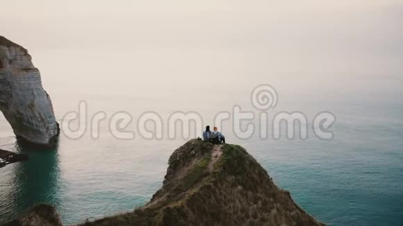 无人驾驶飞机俯视着坐在著名的诺曼底海岸悬崖顶上观看日落海景的浪漫夫妇视频的预览图