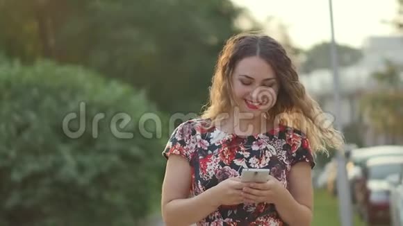 白人迷人的年轻卷发女人在城市公园散步时在智能手机上写短信女人走路视频的预览图