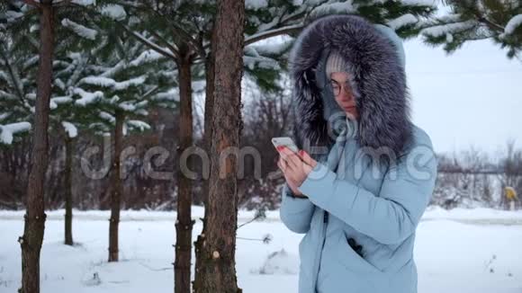 风帽里的小女孩站在一个冬天的松树林里正在找电话里的东西视频的预览图