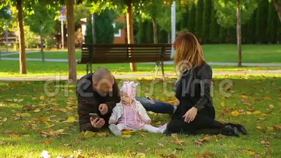 令人惊奇的一对夫妇和一个女儿坐在公园的草地上秋天的树叶是黄色的视频的预览图