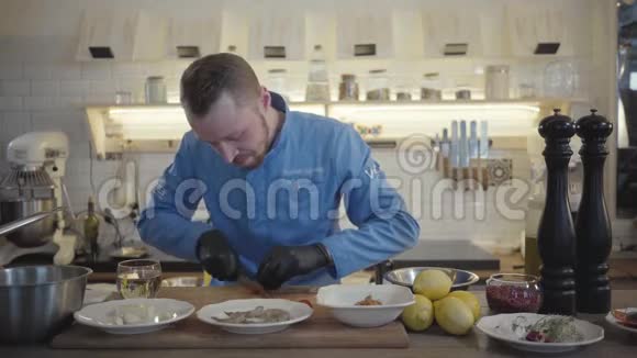 金发厨师穿着餐厅制服戴着黑色手套站在现代化的厨房里周围有许多厨房用具视频的预览图