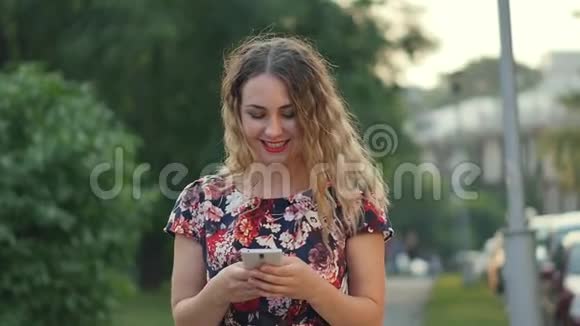 白人迷人的年轻卷发女人在城市公园散步时在智能手机上写短信女人走路视频的预览图