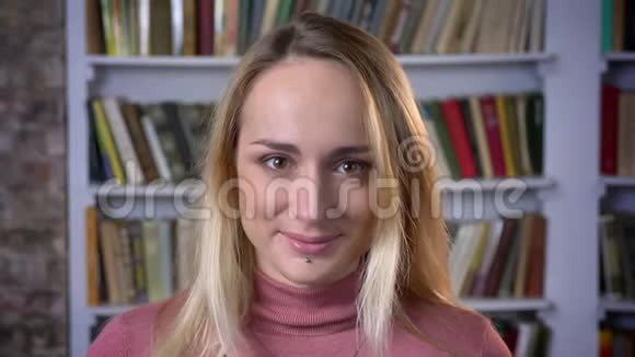 在书架背景上一位古怪的白种人年轻女孩的特写镜头平静地微笑着进入镜头视频的预览图