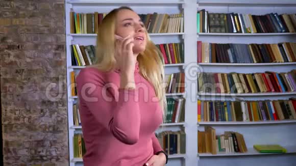 金发白种人年轻女孩的肖像在书架背景上她在手机上愉快地交谈视频的预览图