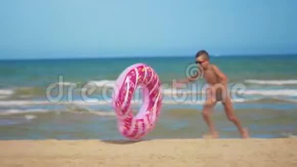 男孩带着一个粉红色的充气甜甜圈沿着海滩跑把它卷在沙滩上贴在大海的背景下视频的预览图